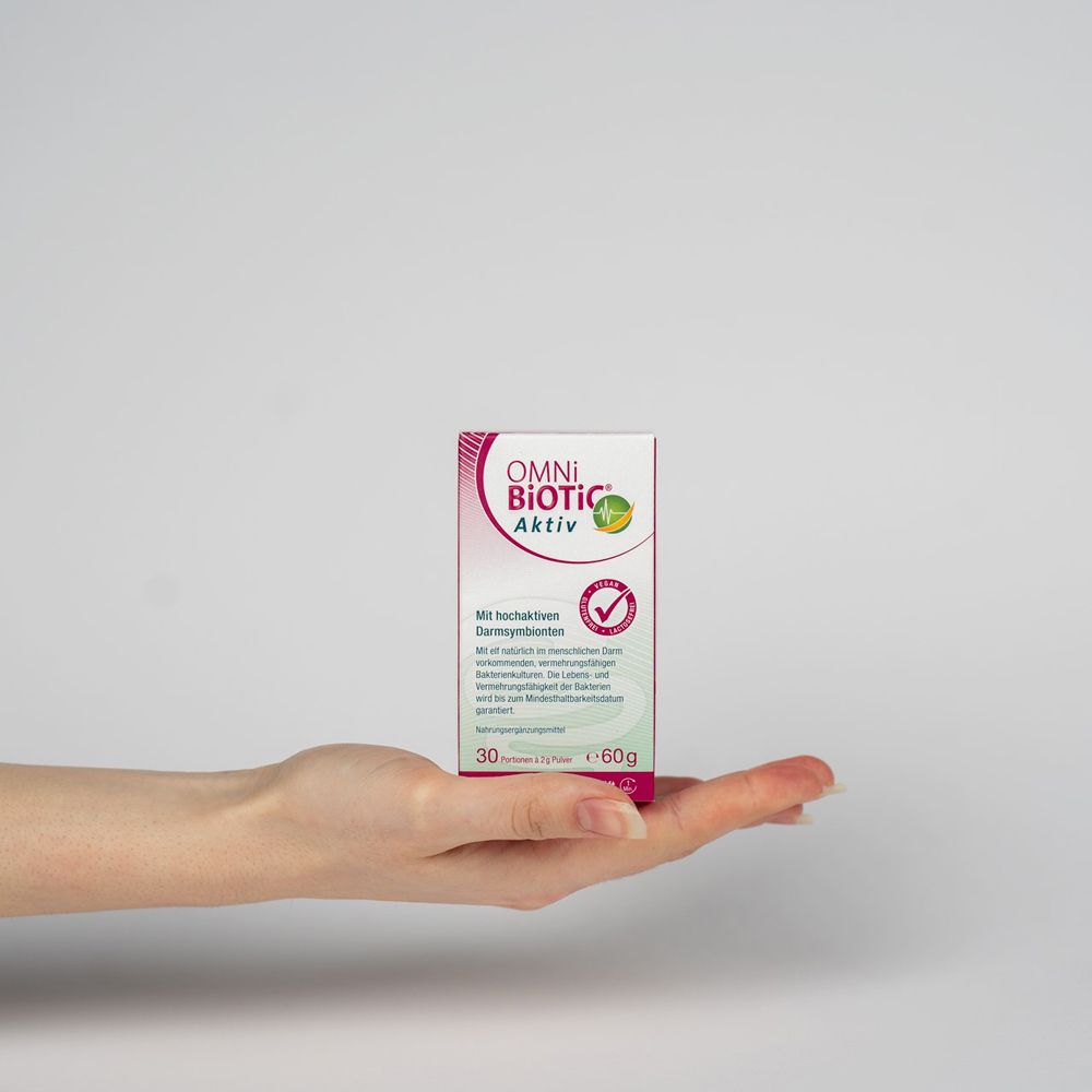 Main tenant une boîte OMNi BiOTiC® Aktiv (60g) pour comparaison de taille. Testé scientifiquement. Végétalien, sans gluten, sans lactose.