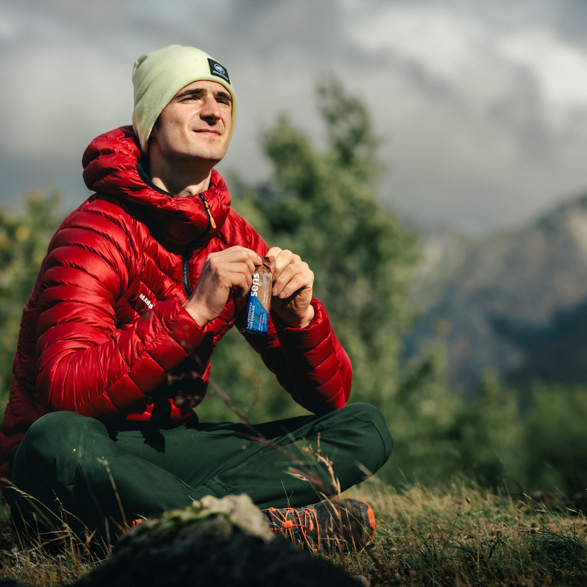 Homme assis dehors mangeant une barre protéinée. Il porte une veste rouge et un bonnet. Montagnes en arrière-plan.
