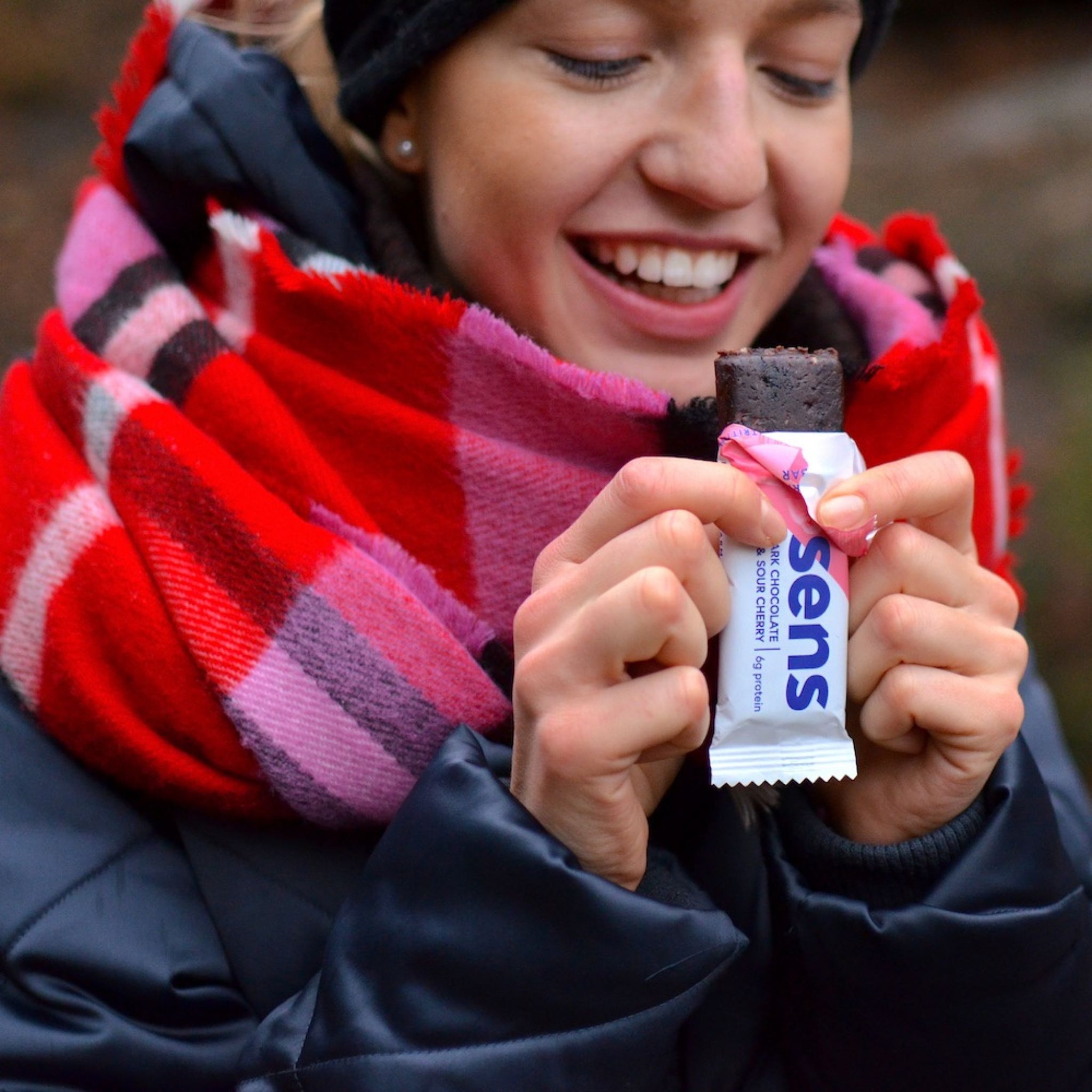 Frau hält einen Proteinriegel in der Hand. Riegel in rosa-weißer Verpackung. Aufschrift: sens. Frau trägt Schal und Mütze.