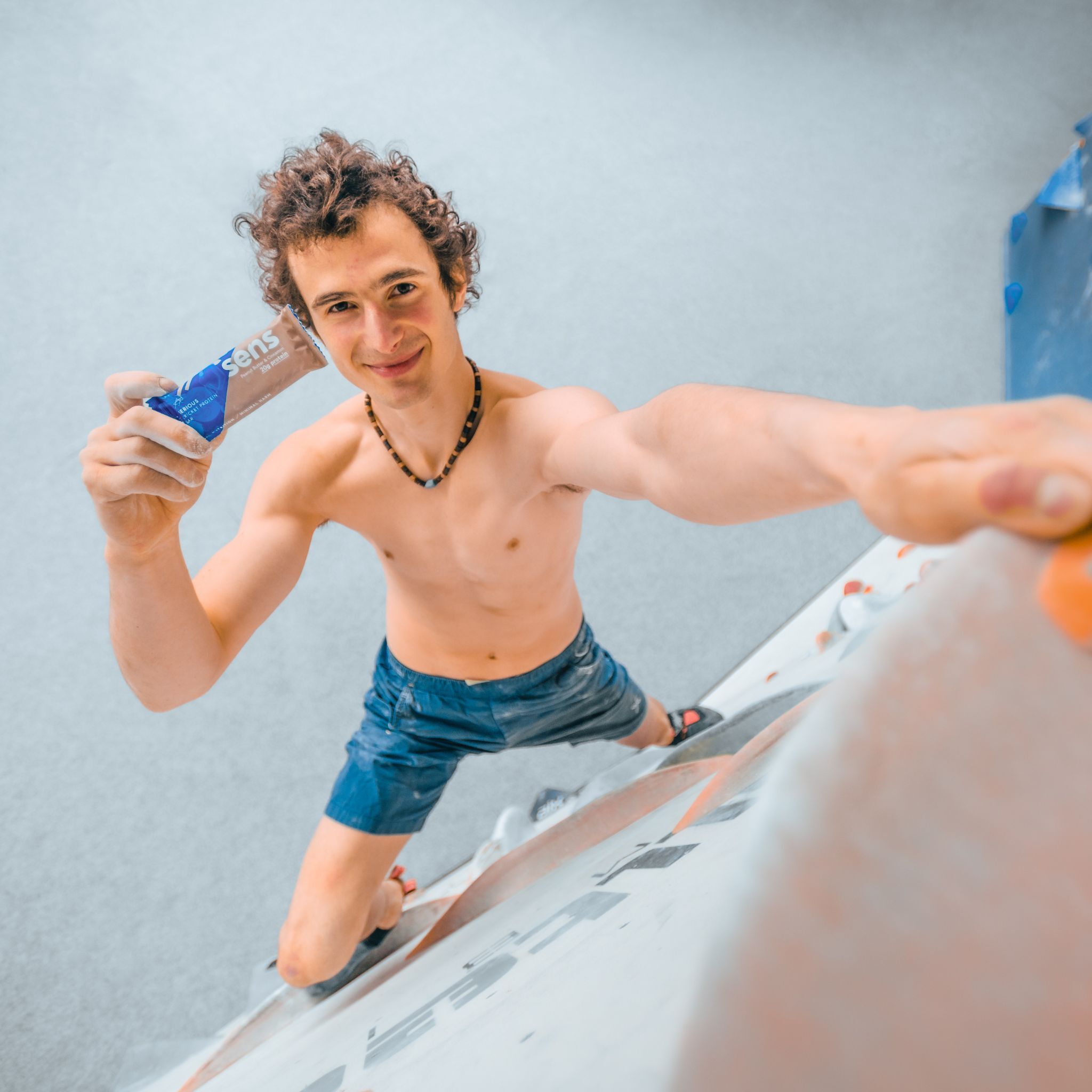 Homme tenant une barre protéinée. Il est pieds nus et porte un short. En arrière-plan, un mur d'escalade.