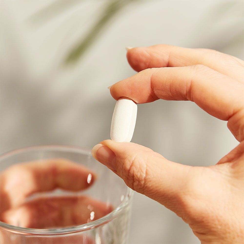 Hand hält eine weiße, ovale Tablette über einem Glas Wasser.