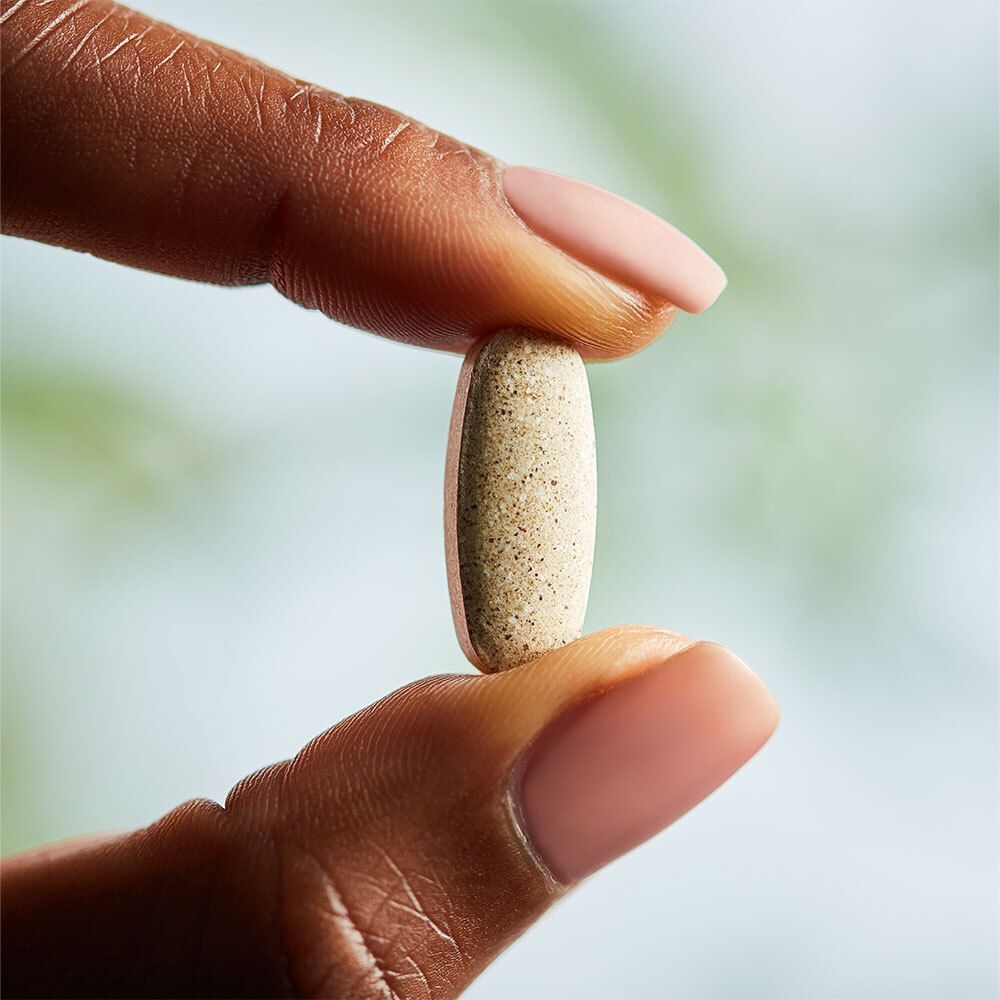 Eine beige Tablette wird zwischen zwei Fingern gehalten. Hintergrund unscharf.