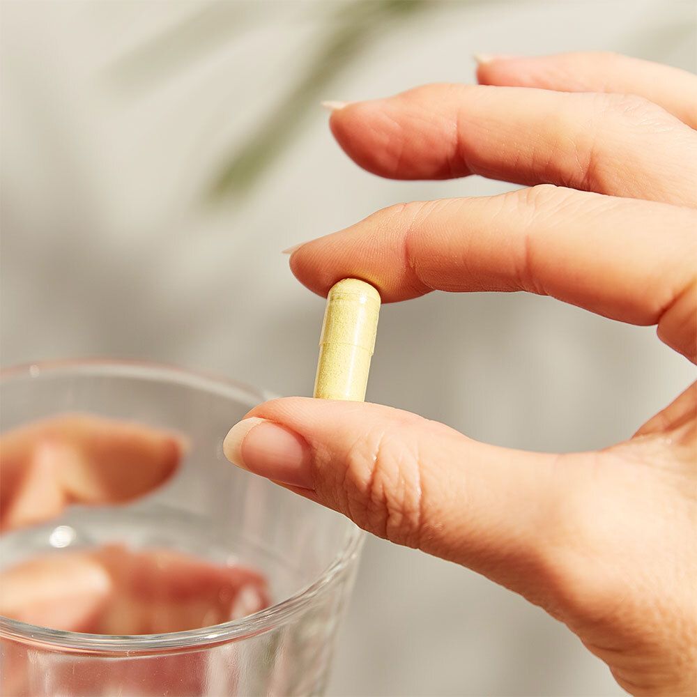 Hand hält eine gelbe Kapsel. Kapsel in der Hand, vor einem Glas Wasser.