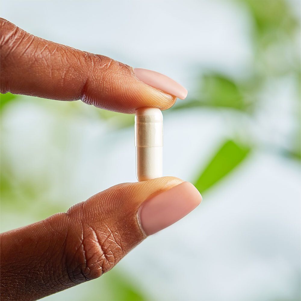 Une main tient une gélule. La gélule est blanche et translucide. Fond : feuillage vert.