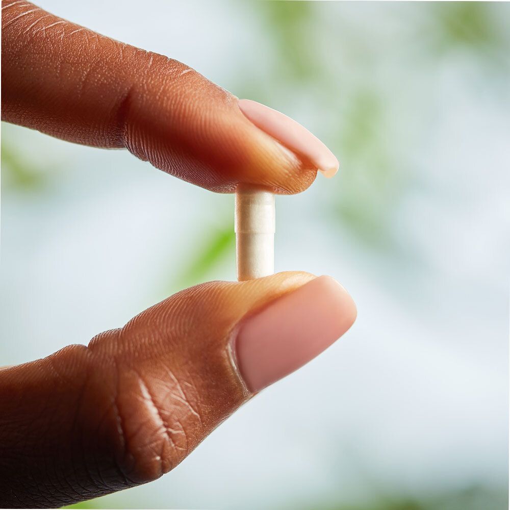 Une gélule entre les doigts. La gélule est blanche et allongée.