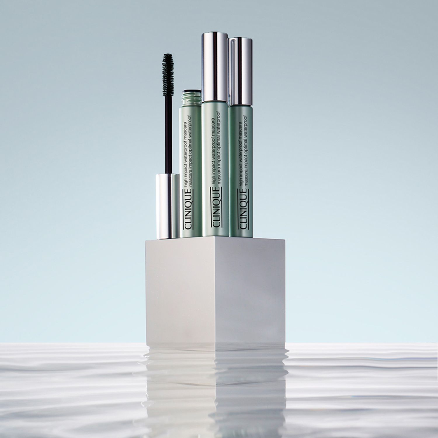 Drei Clinique High Impact Mascara-Tuben auf einem weißen Podest. Wasser im Hintergrund.