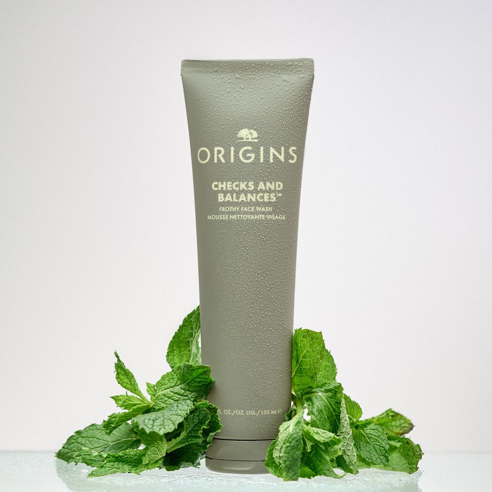 Graue Tube mit Produktnamen und Minzblättern. Text: ORIGINS, CHECKS AND BALANCES, Frothy Face Wash.