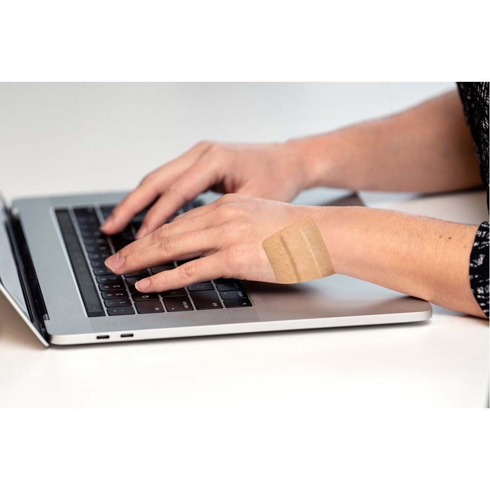 Person tippt auf Laptop, Pflaster am Handgelenk. Helle Oberfläche.