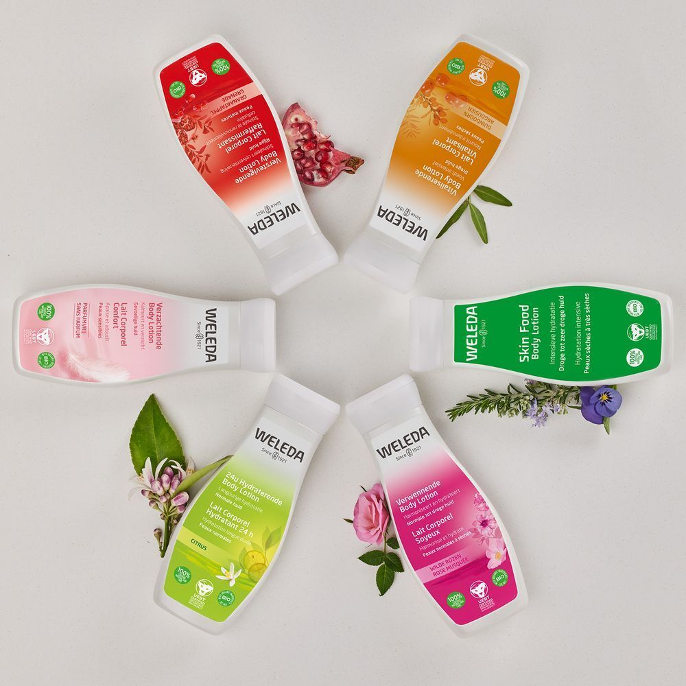 Sechs Weleda-Körperlotion-Flaschen in verschiedenen Farben, angeordnet in einem Kreis. Blumen und Blätter umgeben sie.