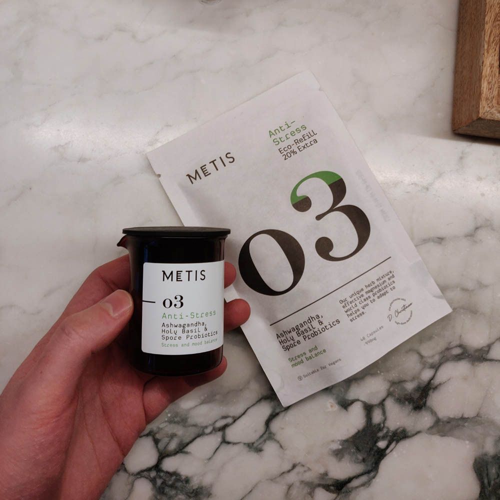 Hand hält eine schwarze Dose und einen weißen Beutel. Dose mit METIS-Logo und "O3"-Aufdruck. Beutel: Anti-Stress, Eco-Refill, 20% Extra.