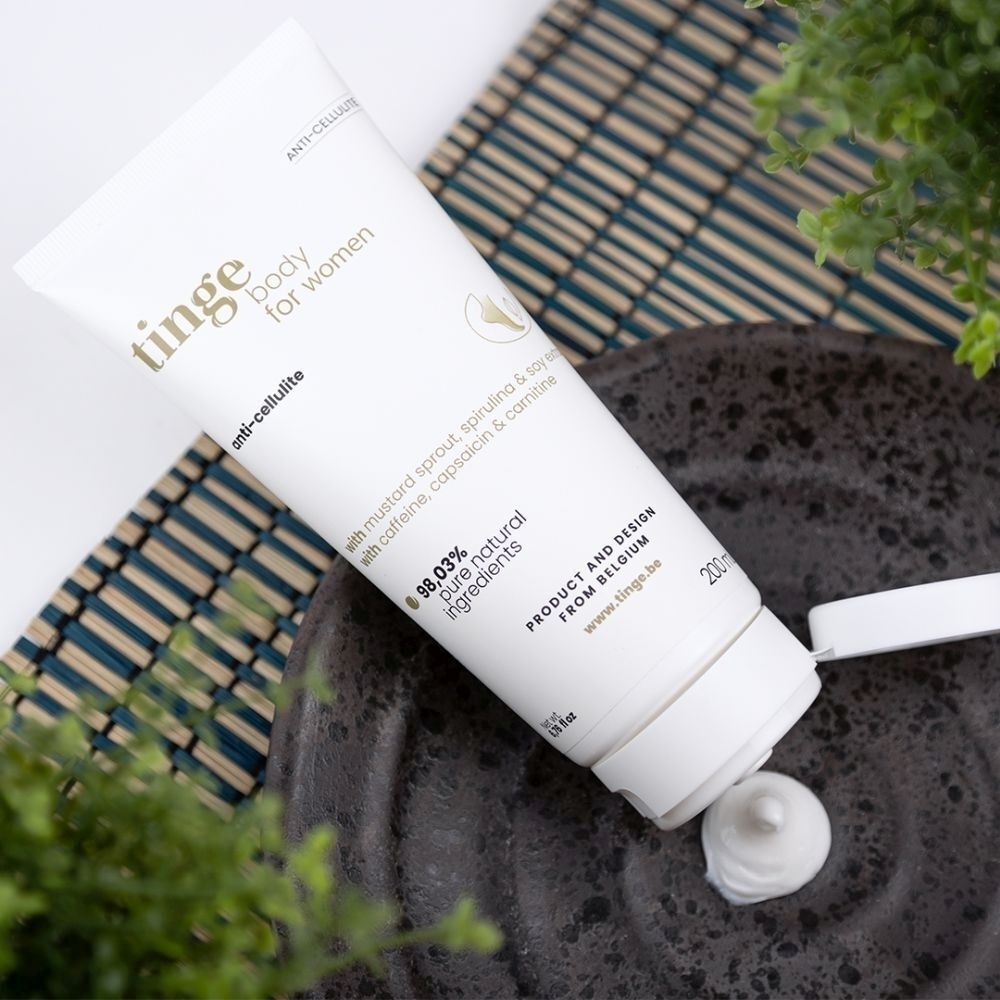Tube de crème anti-cellulite Tinge avec échantillon de produit sur une assiette, entourée de plantes.