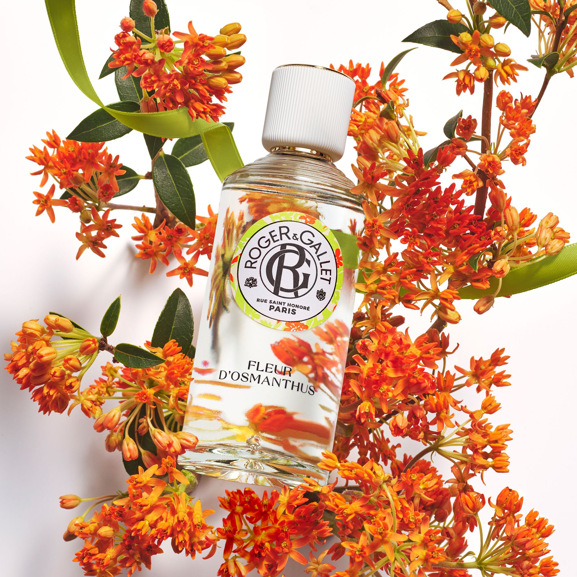 Flacon avec bouchon blanc, étiquette avec logo et nom du produit, entouré de fleurs et feuilles oranges.