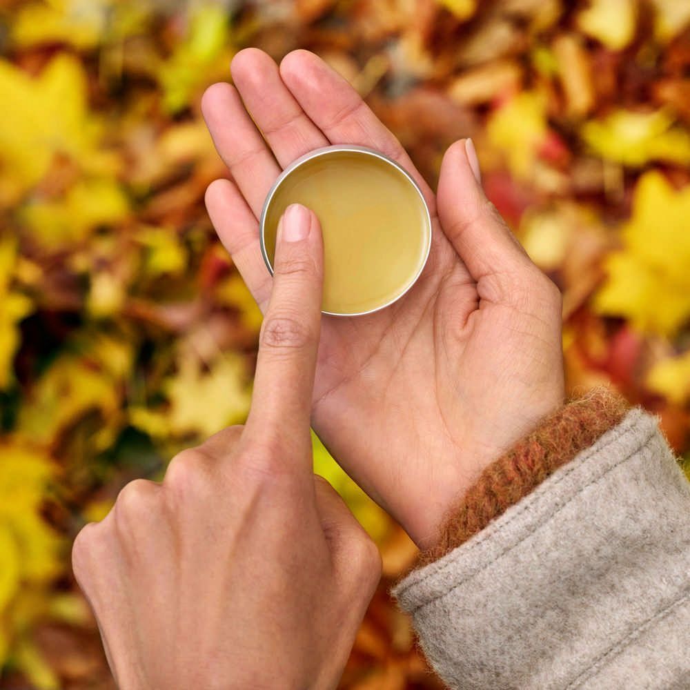 Hände halten eine offene Dose Balsam. Ein Finger berührt den Balsam. Hintergrund: Herbstlaub.