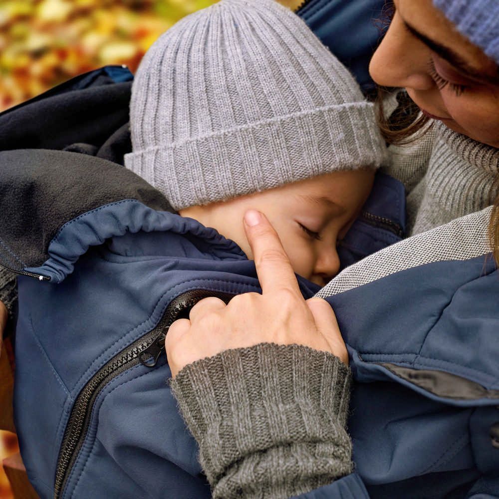 Kind in blauem Anzug, Mütze. Eine Hand berührt sanft das Gesicht des Kindes. Mutter im Hintergrund.