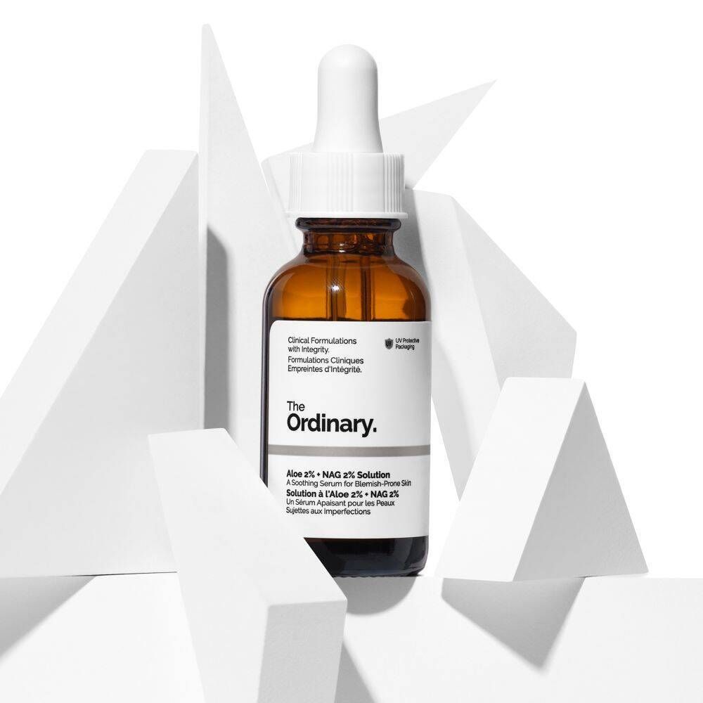 Braune Glasflasche mit Pipette, umgeben von weißen geometrischen Formen. Aufschrift: The Ordinary Aloe 2% + NAG 2% Solution.