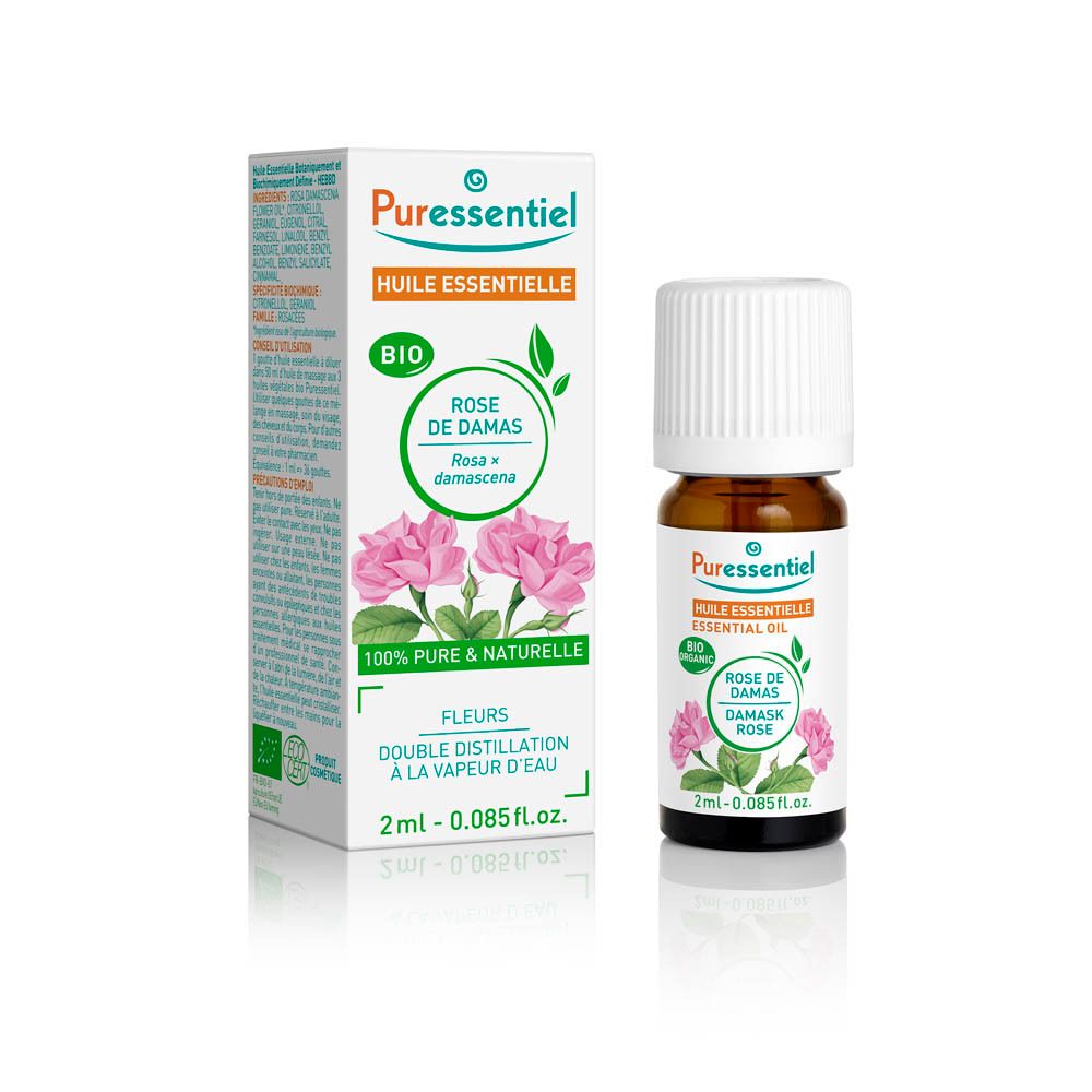Eine kleine Braunglasflasche mit weißem Schraubverschluss, gefüllt mit ätherischem Rosenöl. Daneben eine geöffnete Pappschachtel mit Rosenabbildungen.