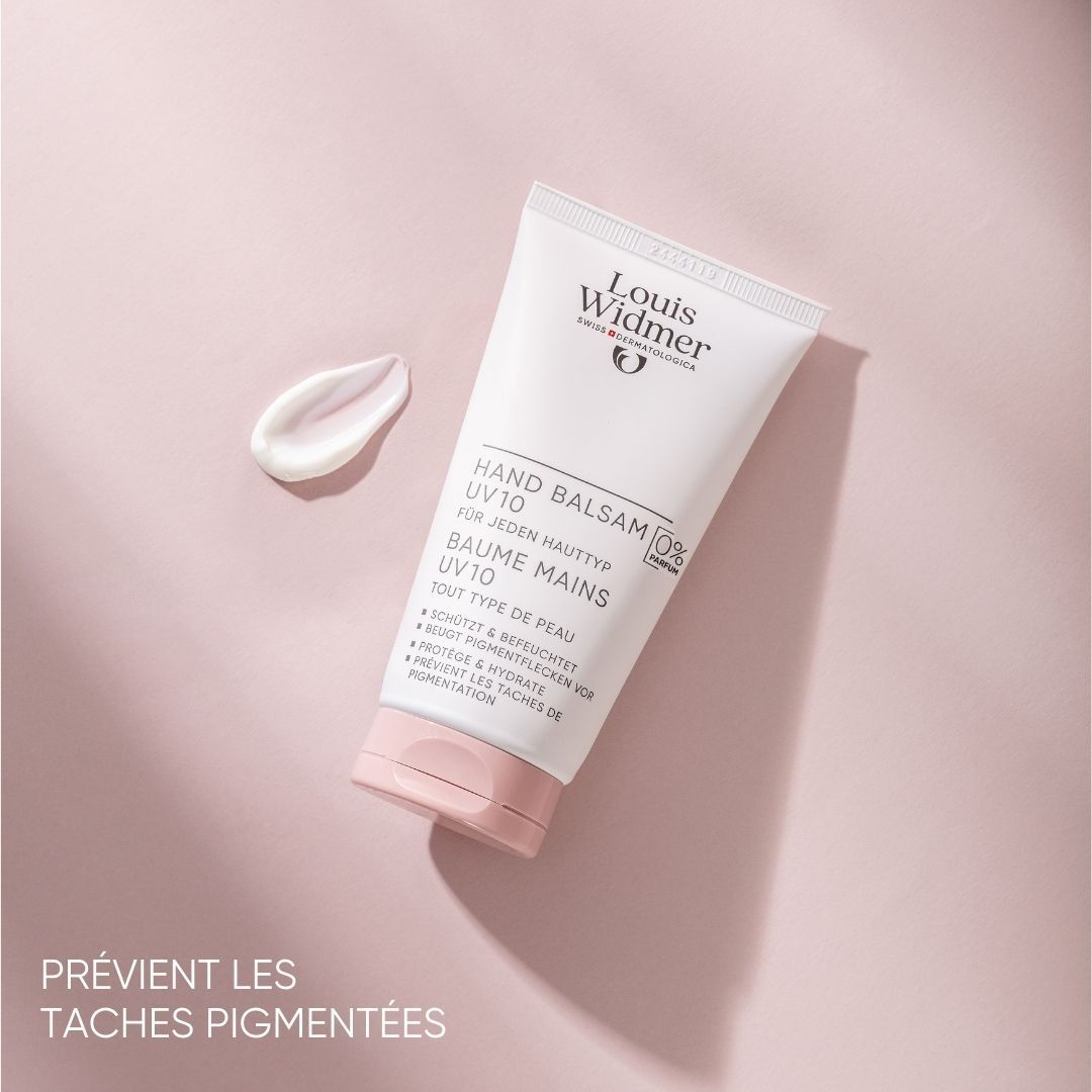 Tube blanc avec nom et logo du produit. Texte: Hand Balsam UV10, 0% Parfum. Sur fond rose. Goutte de crème à côté.
