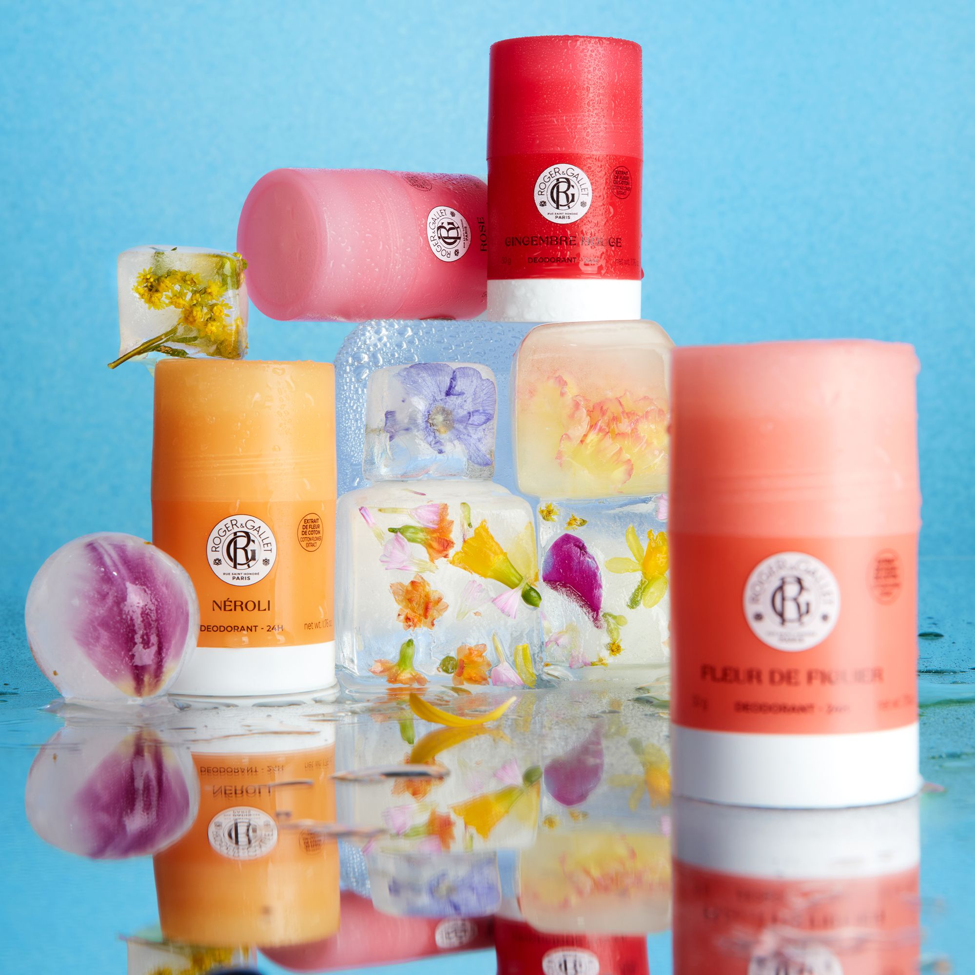 Déodorants Roger & Gallet Néroli. Cubes de glace avec fleurs. Emballages roses, oranges et rouges. Sur fond bleu.