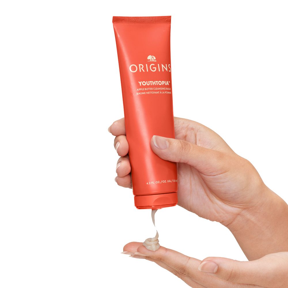 Main tenant un tube rouge. Le produit est appliqué sur le doigt. Inscription: ORIGINS YOUTHTOPIA.