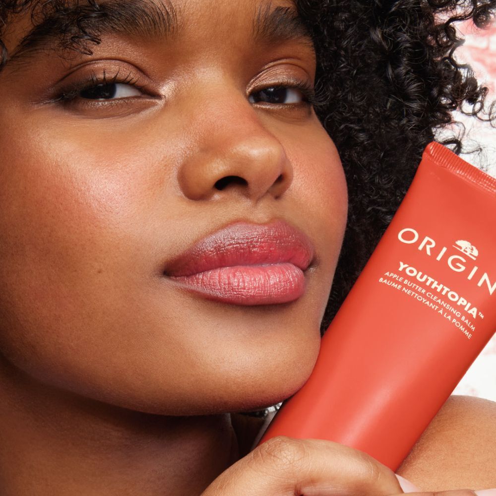 Femme tenant un tube rouge. Inscription: ORIGINS YOUTHTOPIA Apple Butter Cleansing Balm.