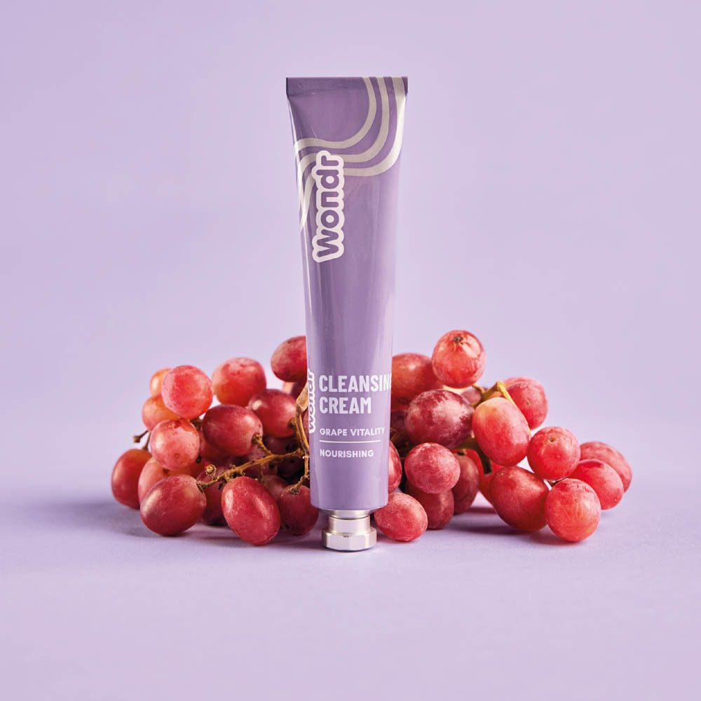 Lila Tube mit "Wondr"-Logo und "Cleansing Cream Grape Vitality" Schriftzug. Liegt auf Trauben. Auf lila Hintergrund.