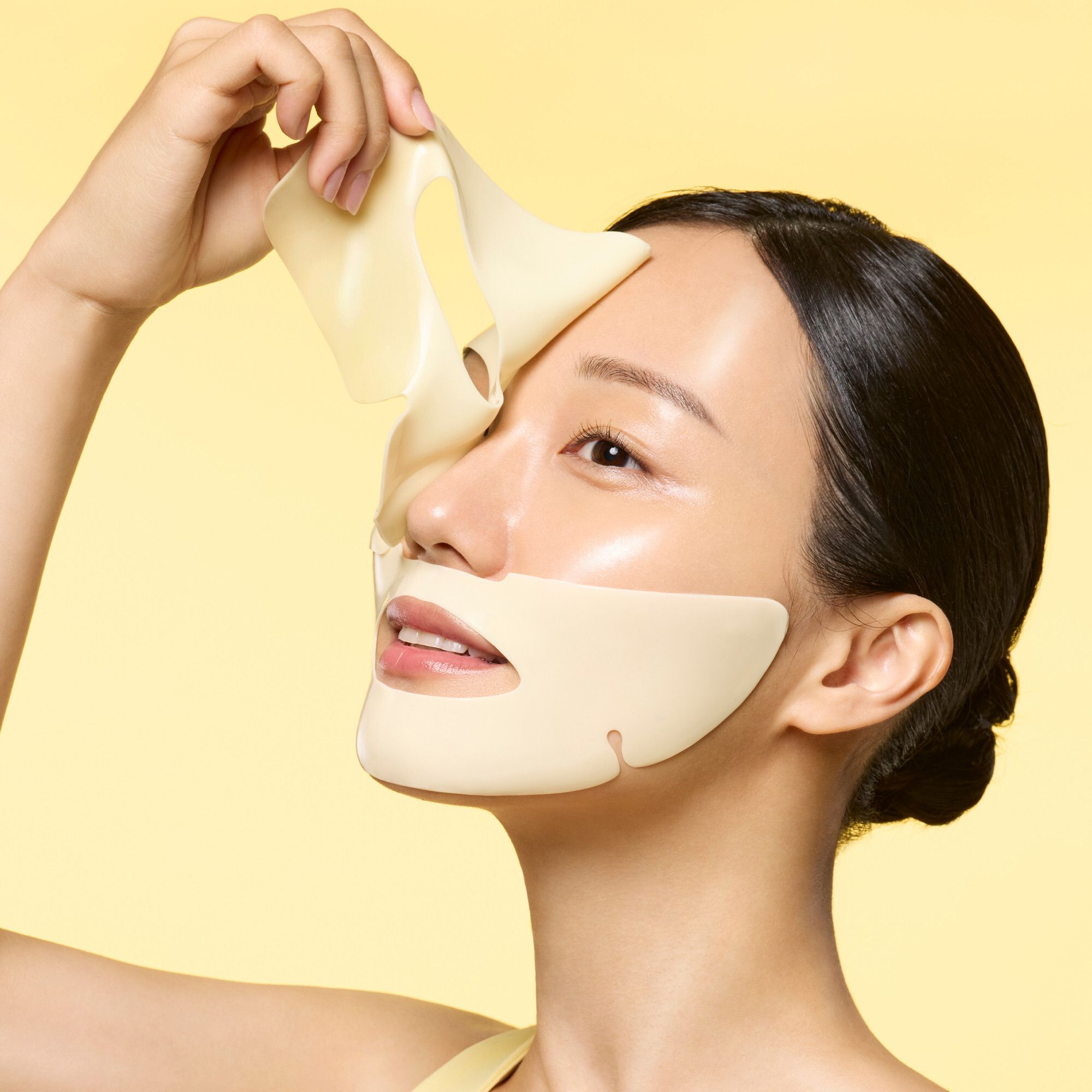 Une femme tient une feuille de masque jaune de Dr.Jart+ Cryo Rubber™ Brightening Mask contre son visage.
