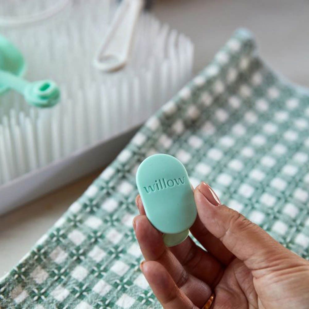 Une main tenant une valve Elvie Wave vert menthe avec logo 'willow'. Un égouttoir et une serviette en arrière-plan.