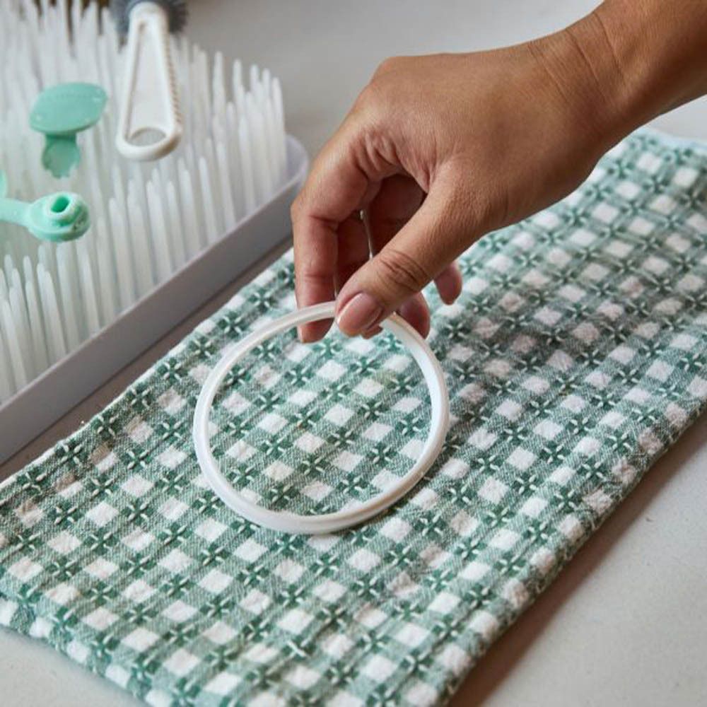 Eine Hand hält einen weißen Silikonring über einem Handtuch. Im Hintergrund ist ein Abtropfgestell mit Babyflaschenzubehör zu sehen.