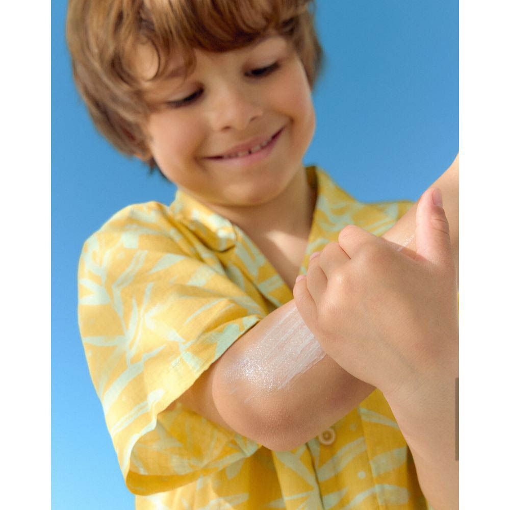 Un enfant reçoit de la crème solaire sur le bras. Chemise jaune.