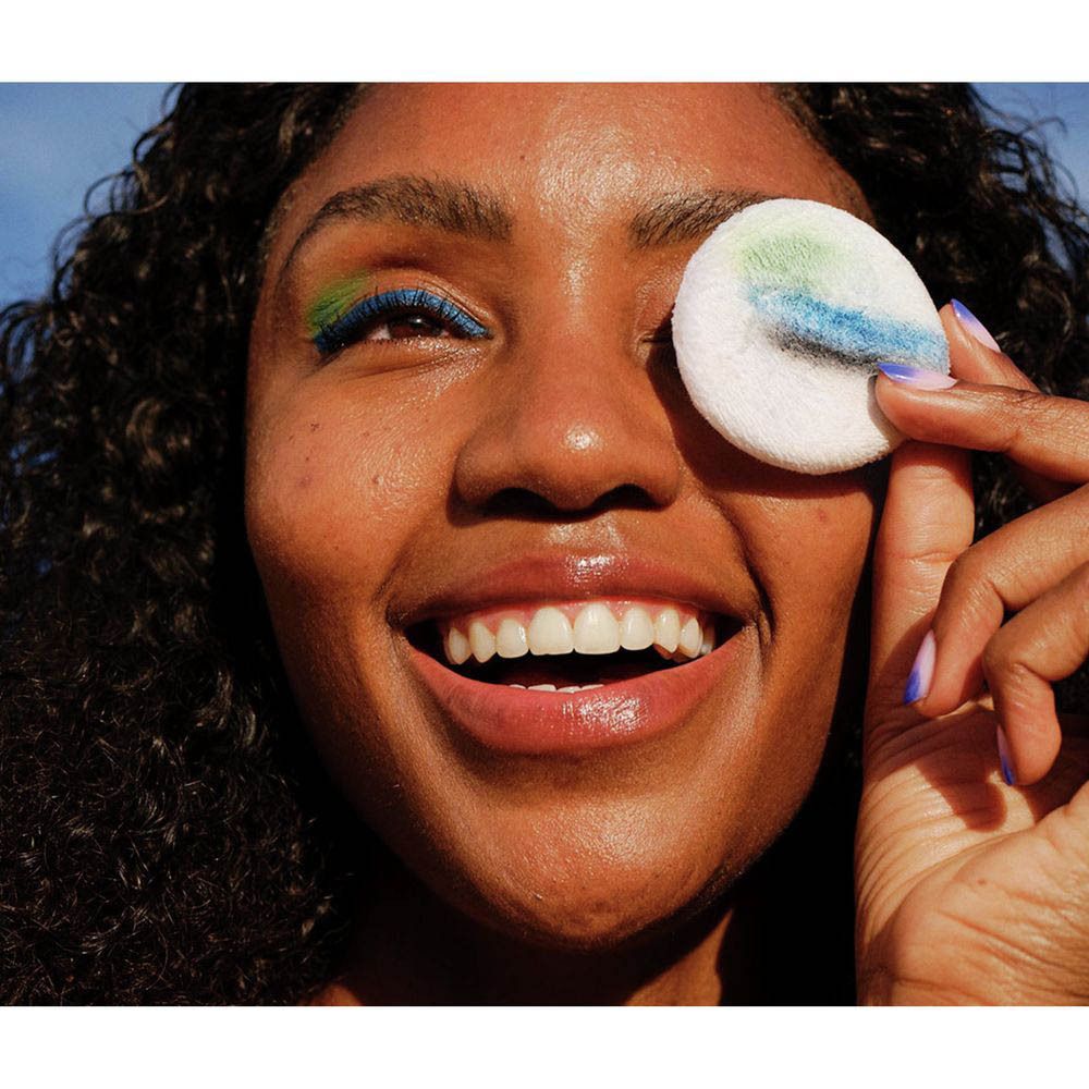Femme avec maquillage coloré pour les yeux tenant un coton démaquillant avec des restes de maquillage devant son œil. Nivea Skin Glow Eau Micellaire.