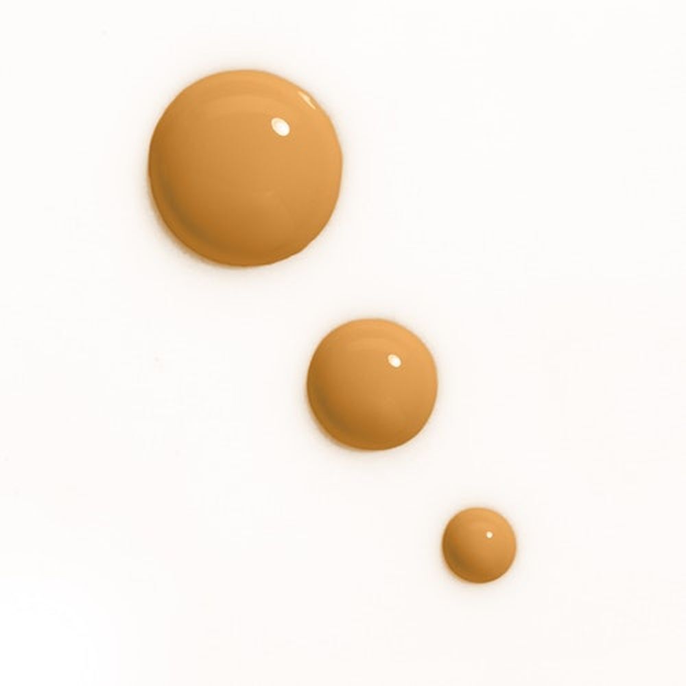 Drei Tropfen Foundation in verschiedenen Größen. Helle, goldbraune Farbe auf weißem Hintergrund.