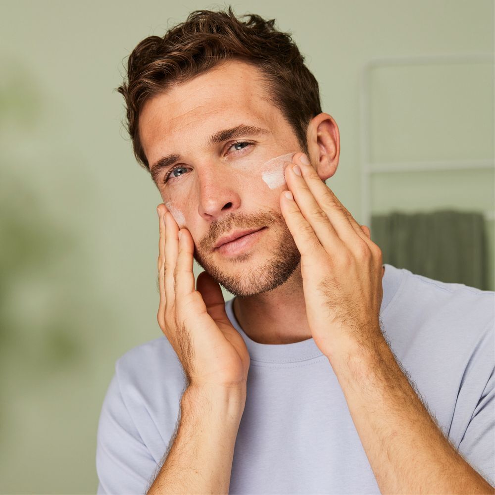Ein Mann trägt Creme auf sein Gesicht auf. Er berührt seine Wangen mit den Händen. Helles Badezimmer im Hintergrund.
