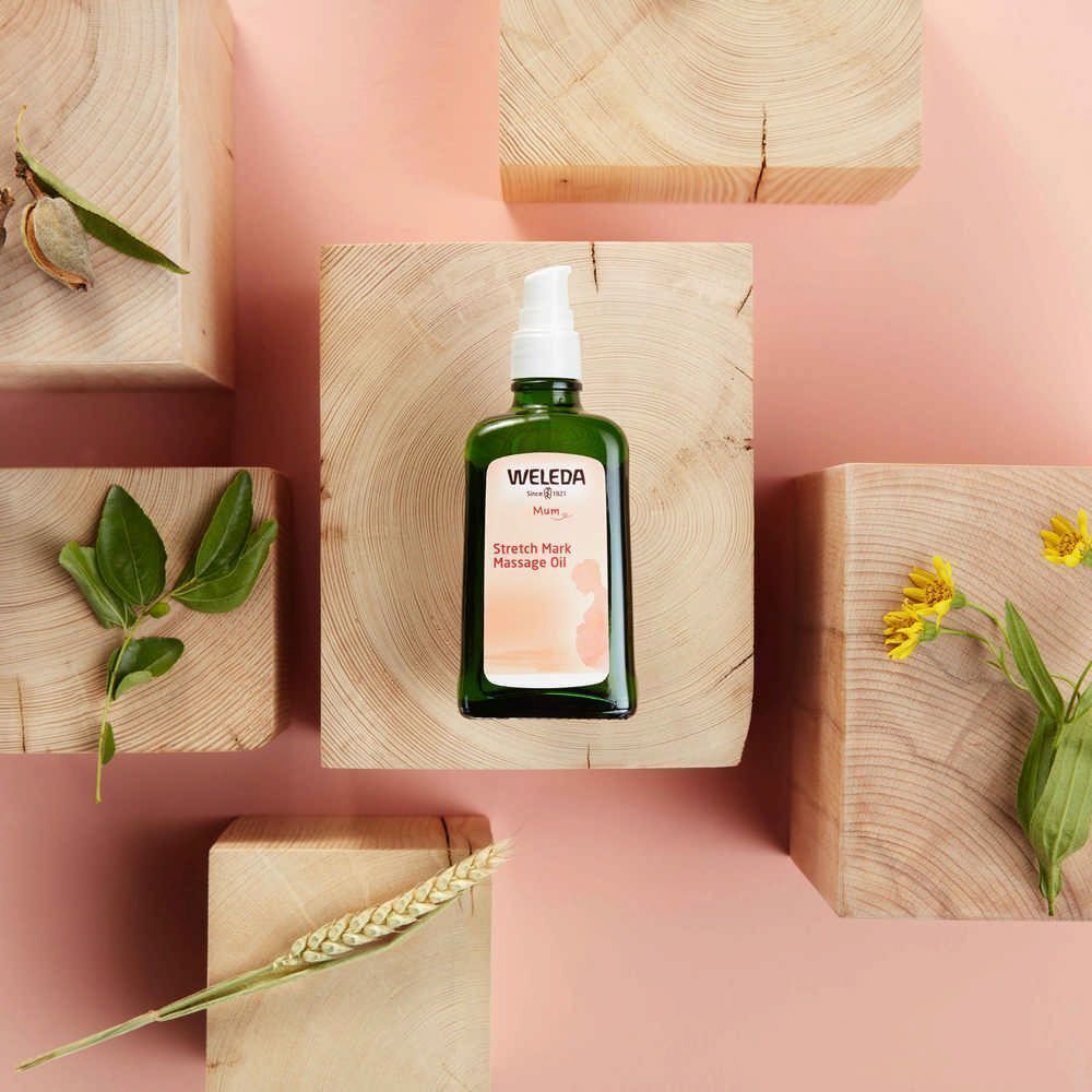 Grüne Flasche Weleda Mama Massageöl auf Holzblöcken. Umgeben von Holz, Blättern und Blüten.