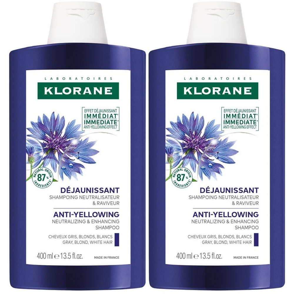 Deux flacons de shampooing Klorane anti-jaunissement. Bouteilles violettes avec bouchons et étiquettes blancs. 87% ingrédients d'origine naturelle.