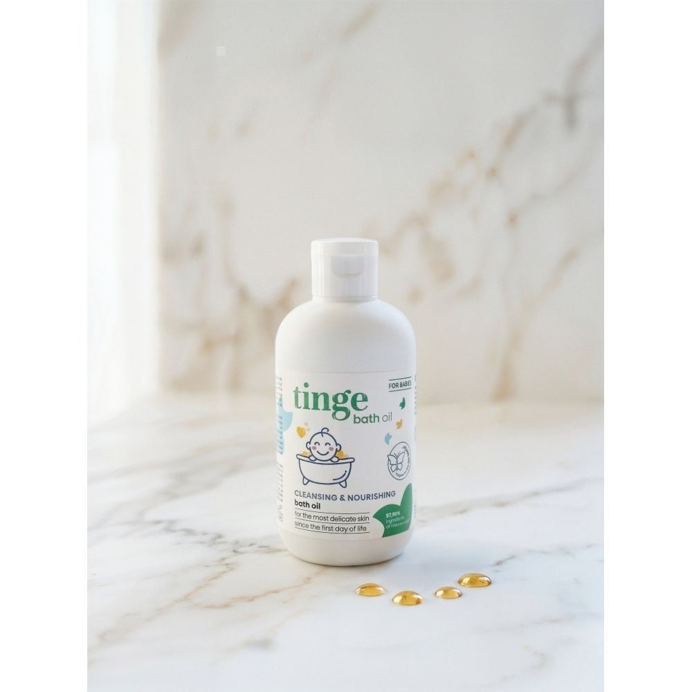 Un flacon d'huile de bain Tinge pour bébés sur une surface en marbre avec quelques gouttes dorées à côté.