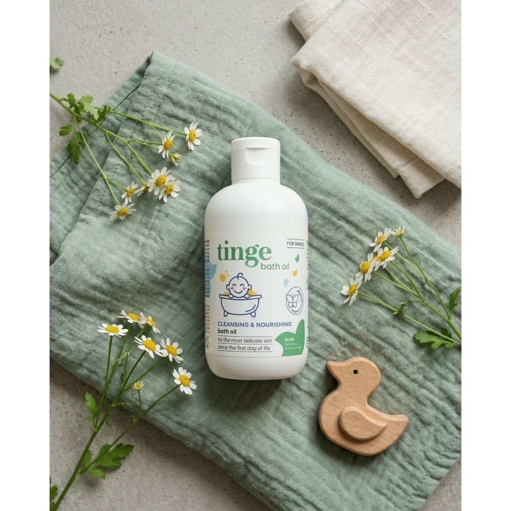 Un flacon d'huile de bain Tinge pour bébés sur un tissu vert, avec de la camomille et un canard en bois.
