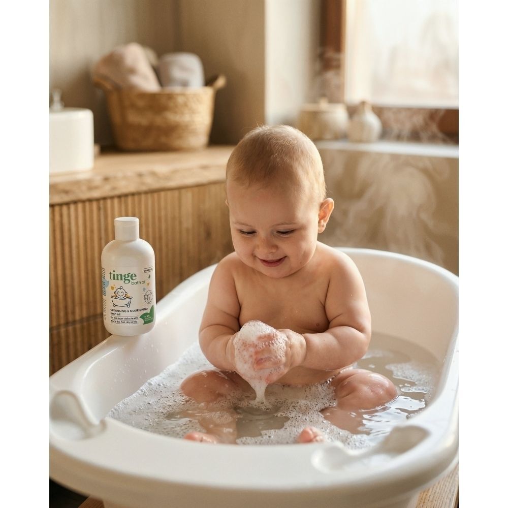 Un bébé assis dans une baignoire moussante à côté d'un flacon d'huile de bain Tinge pour bébés.