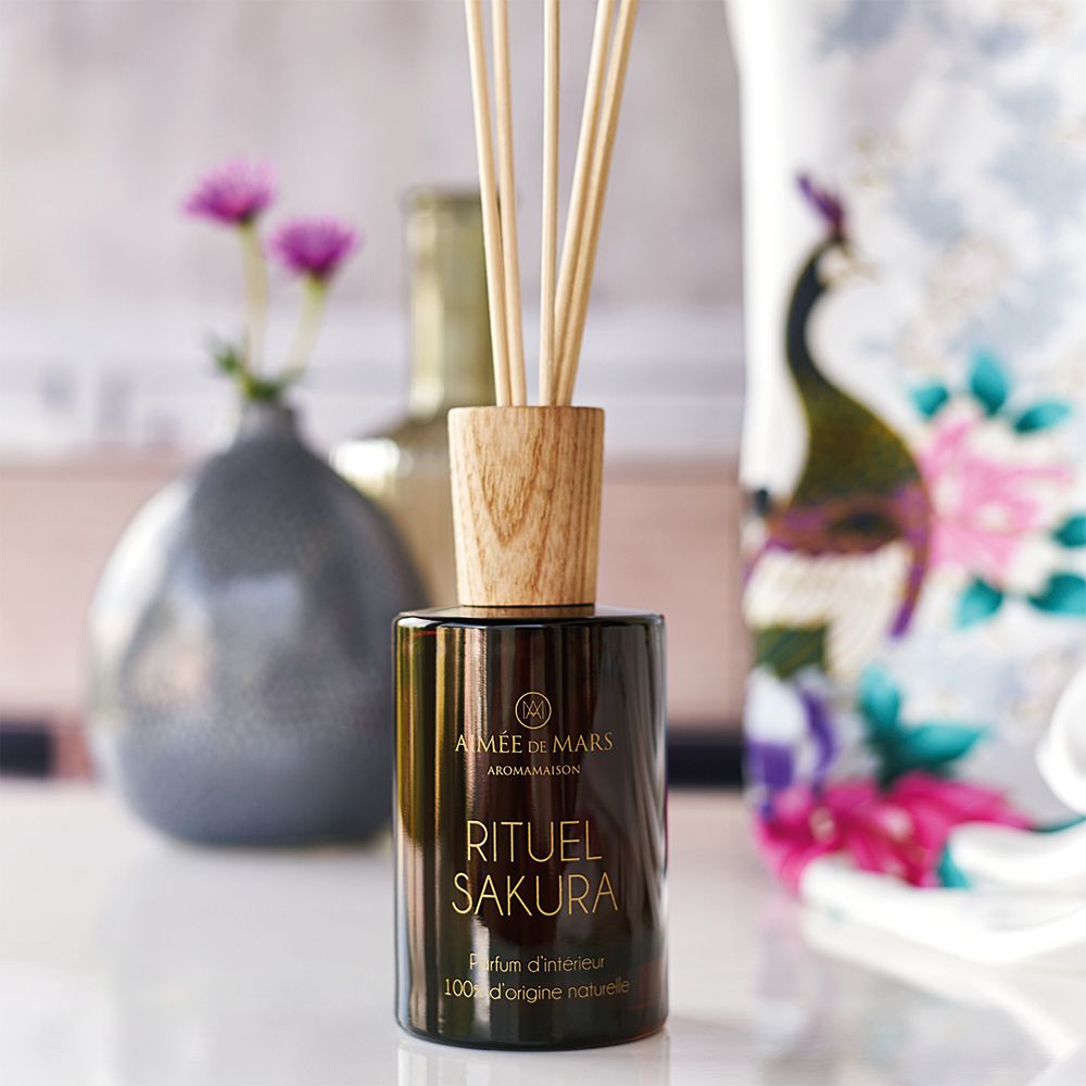 Braune Glasflasche mit Holzdeckel und Stäbchen. Aufschrift: Rituel Sakura. Im Hintergrund Vasen und Blumen.