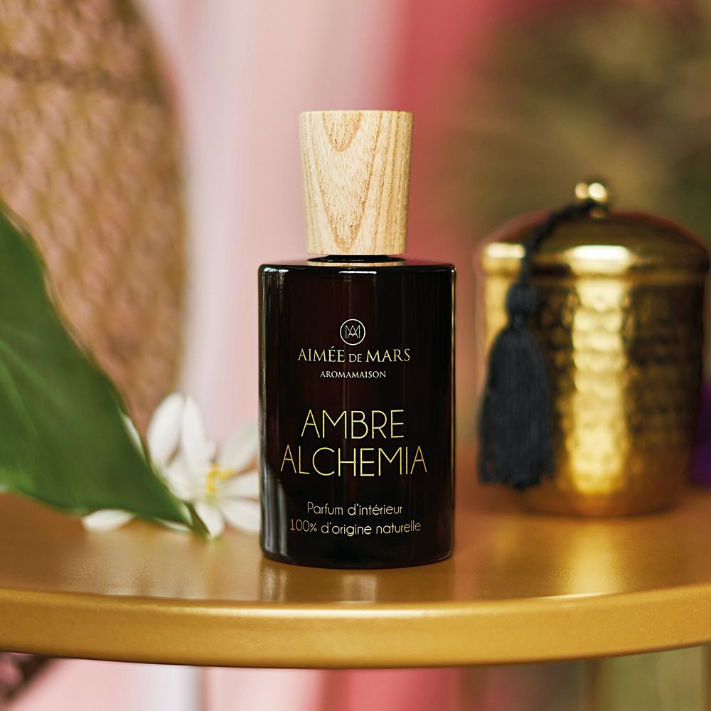 Flacon en verre brun avec bouchon en bois. Inscription: AMBRE ALCHEMIA, Parfum d'intérieur, 100% d'origine naturelle. Sur table dorée.