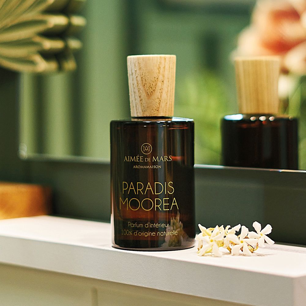 Braune Glasflasche mit Holzdeckel auf einer weißen Ablage. Aufschrift: Aimée de Mars, Paradis Moorea. Weiße Blüten daneben.