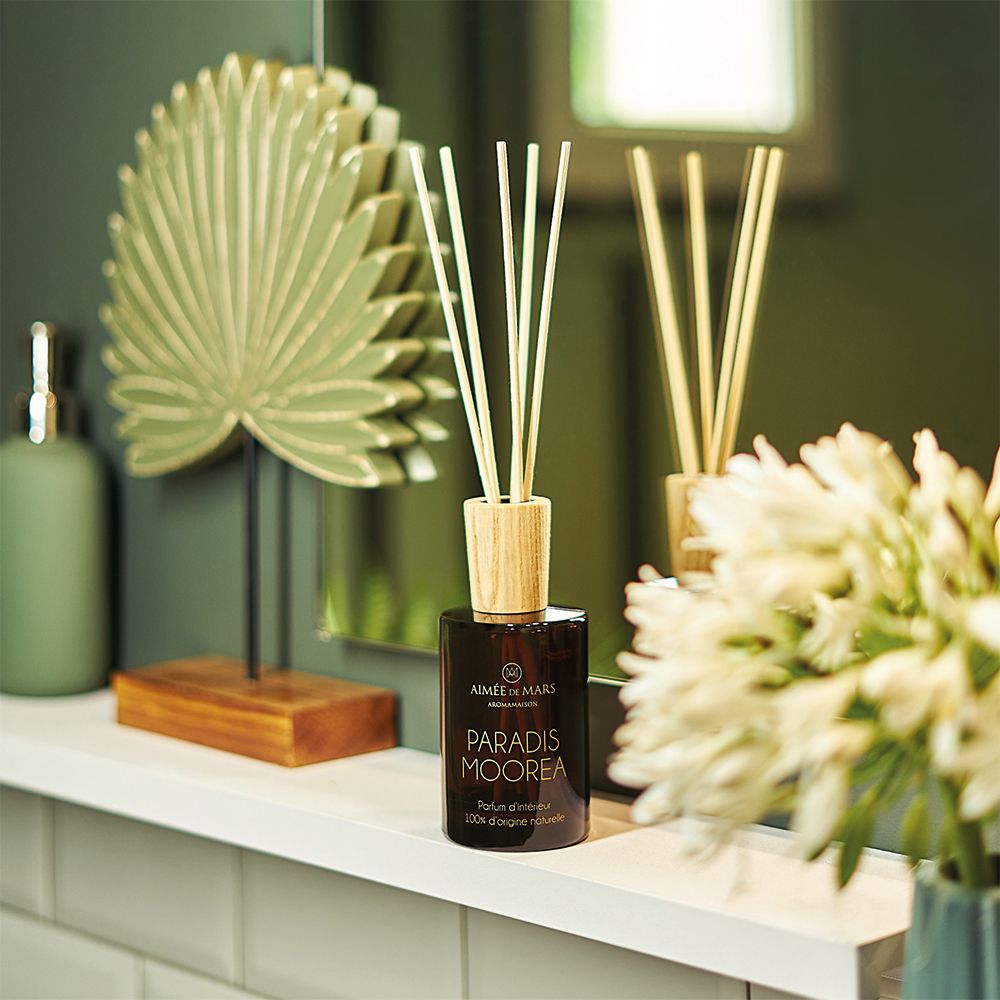 Braune Glasflasche mit Holzdeckel und Holzstäbchen. Auf einer weißen Ablage, neben Dekoration und Spiegel.