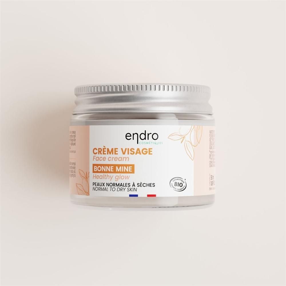 Pot de crème avec couvercle argenté. Étiquette avec nom du produit, marque et label bio. Texte: Crème Visage, Bonne Mine.