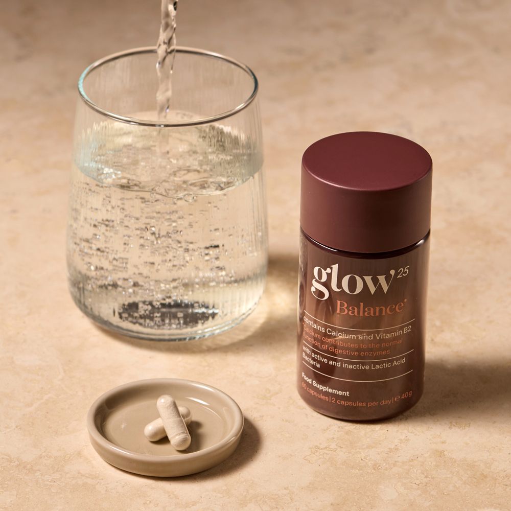 Glow25 Balance Dose neben einem Glas Wasser und zwei Kapseln auf einem kleinen Teller.