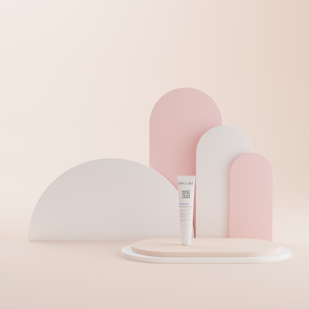 Tube blanc DECLARÉ Eye Contour Firming Cream sur socle. Fond avec formes roses et blanches.