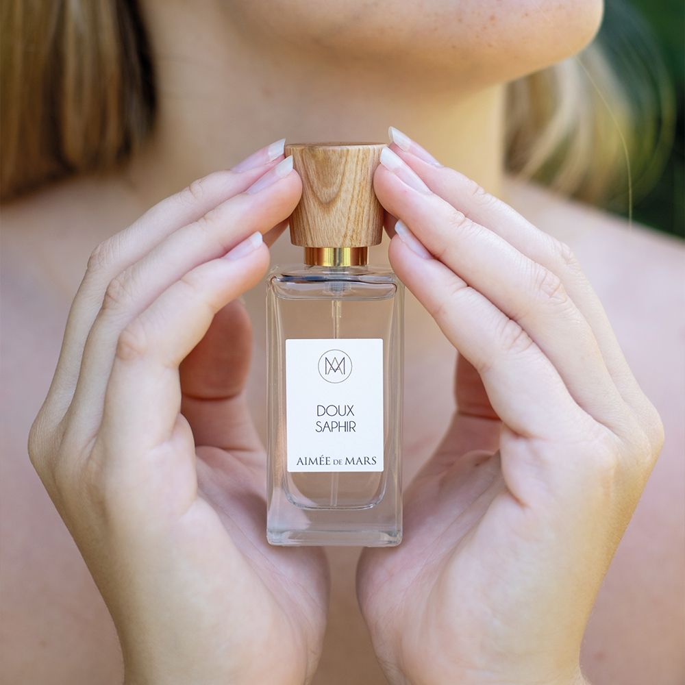 Hände halten einen Parfümflakon mit Holzverschluss. Auf dem Etikett steht "Doux Saphir" und "Aimée de Mars".
