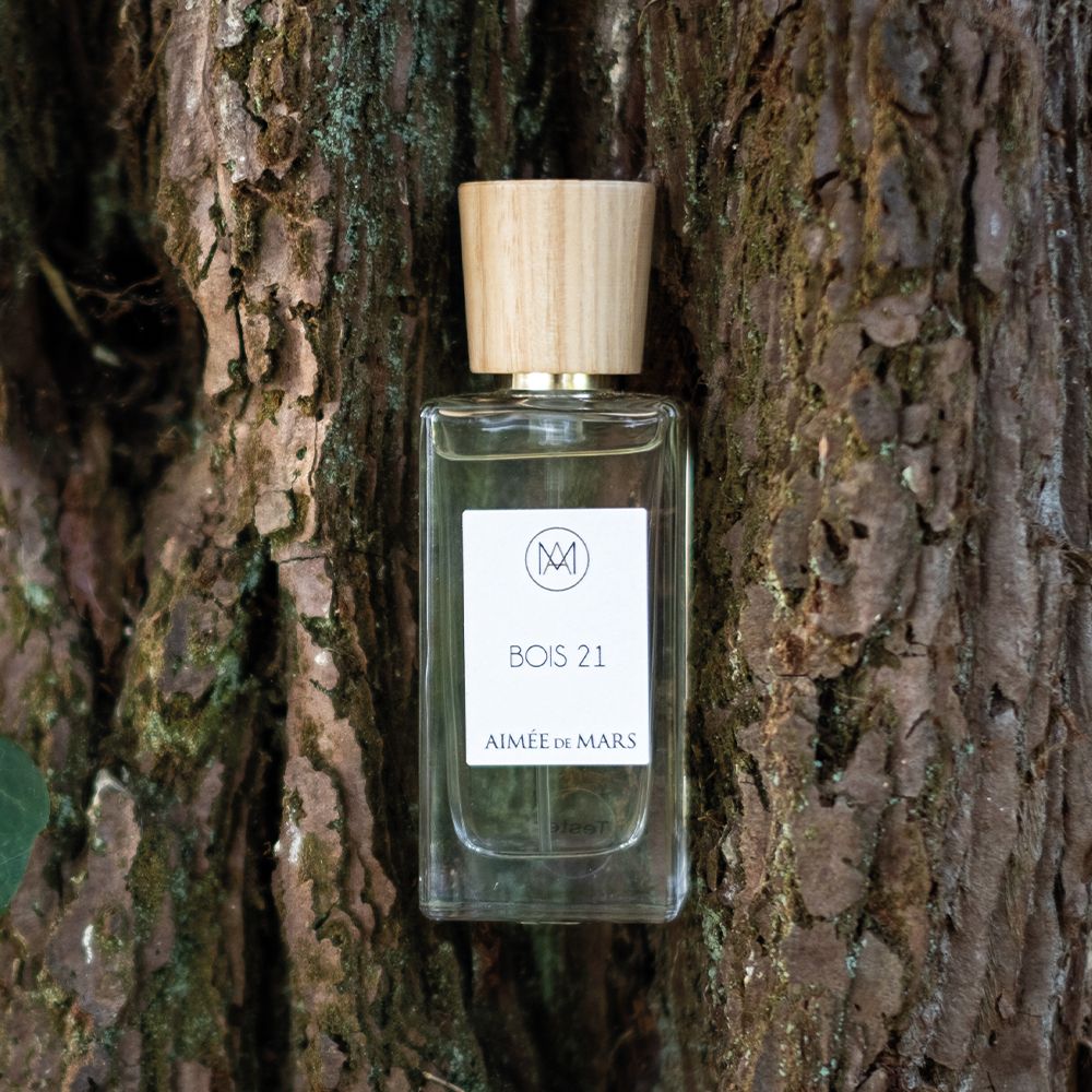 Glasflakon mit Holzdeckel vor Baumrinde. Aufkleber mit Logo, Schriftzug „Bois 21“ und „Aimée de Mars“. Flüssigkeit sichtbar.