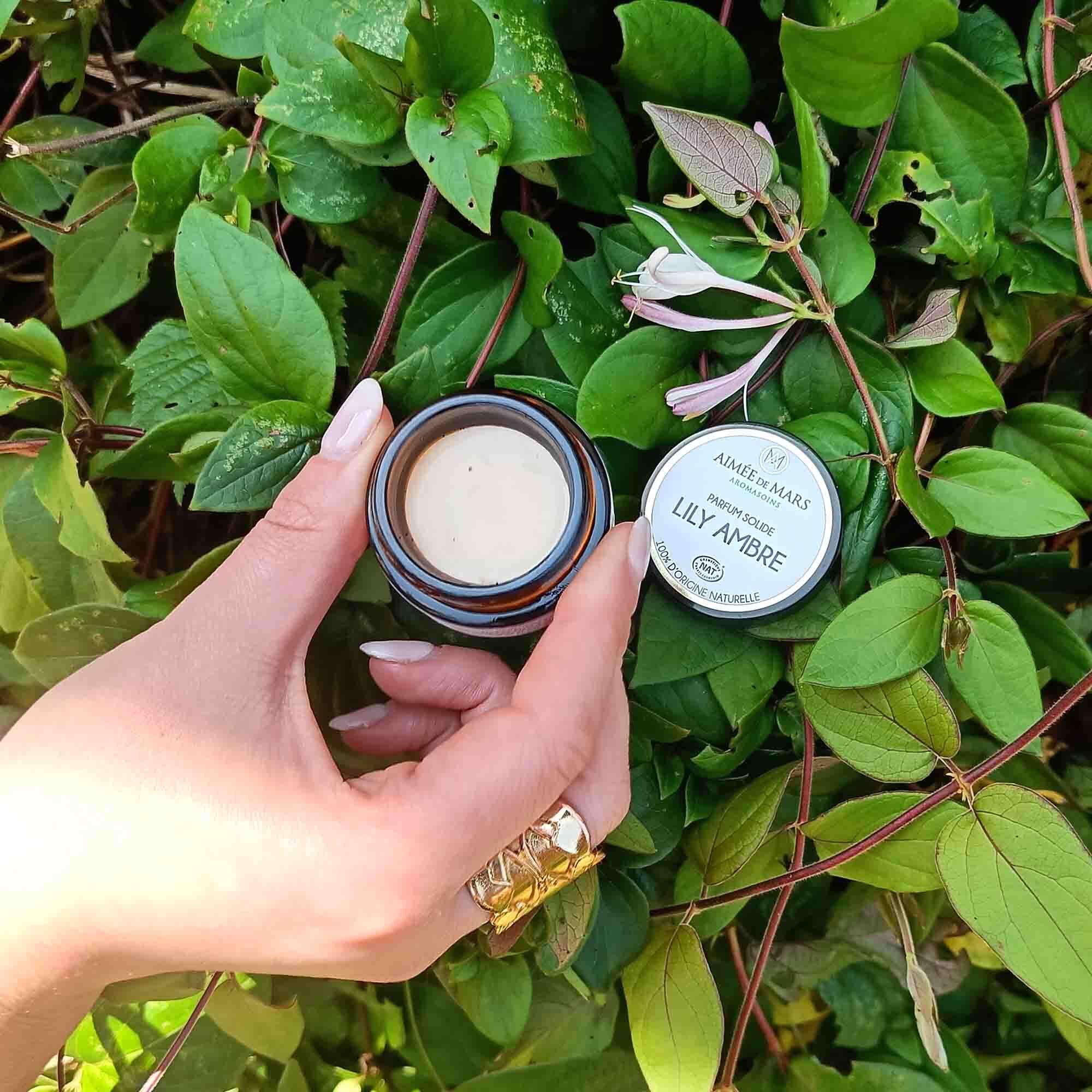 Hand hält ein geöffnetes braunes Glas mit cremefarbenem Inhalt und einen Deckel. Aufschrift "LILY AMBRE".