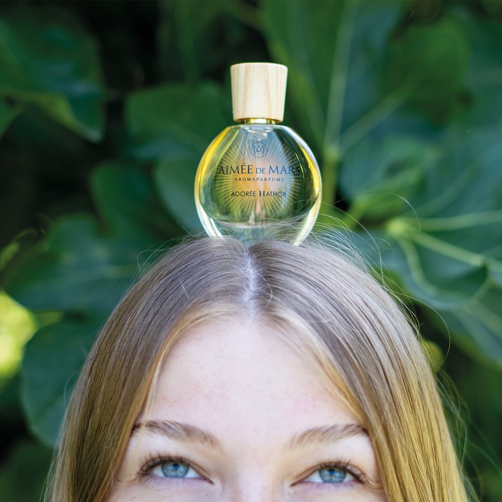 Frau mit Flakon auf dem Kopf. Flakon: rund, Glas, Holzverschluss. Aufschrift: AIMÉE DE MARS, ADORÉE HATHOR.