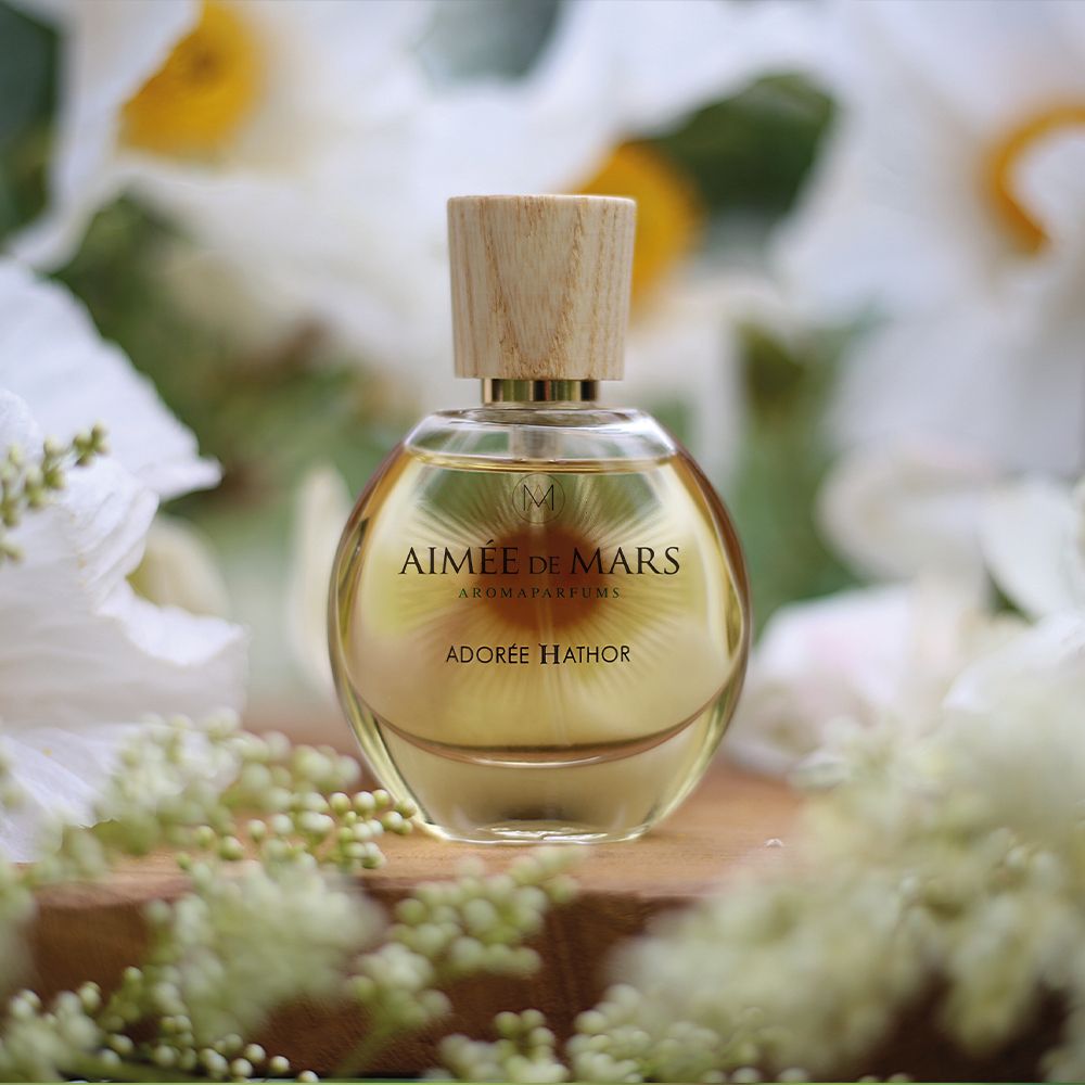 Flakon auf Holzbrett, umgeben von Blüten. Flakon: rund, Glas, Holzverschluss. Aufschrift: AIMÉE DE MARS, ADORÉE HATHOR.