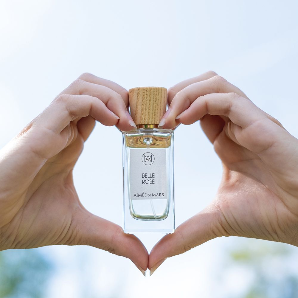 Aimée de Mars Belle Rose Eau de Parfum Flakon, gehalten von Händen in Herzform. Rechteckige Glasflasche mit Holzverschluss.